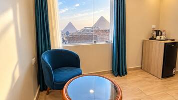 Habitación de hotel con vista a las pirámides, un sillón azul, una mesita de vidrio y un minibar.