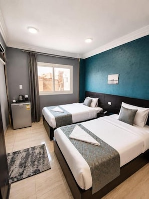 Ein Hotelzimmer mit zwei Betten, einem Schreibtisch, einer kleinen Kühlbox und einem Fenster mit Vorhängen.