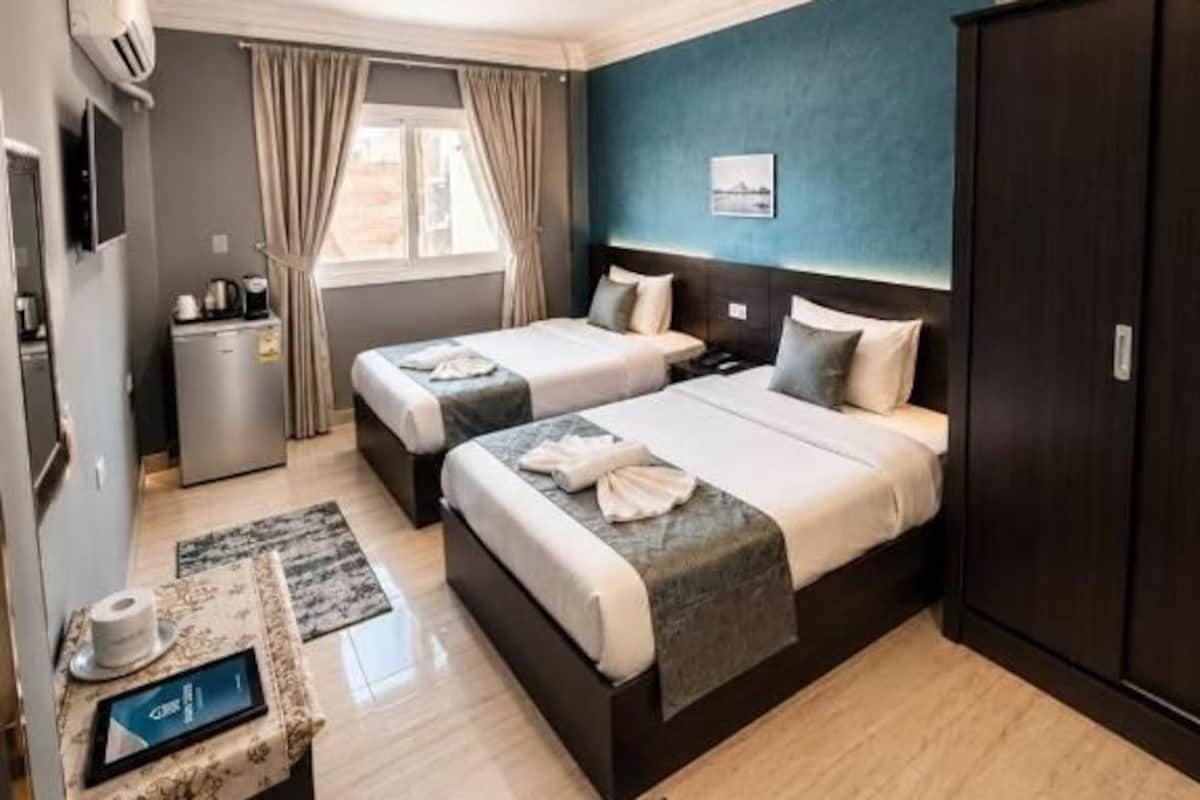 Ein Hotelzimmer mit zwei Betten, einem Schreibtisch mit Computer, einem Kleiderschrank und einem Fenster mit Vorhängen.
