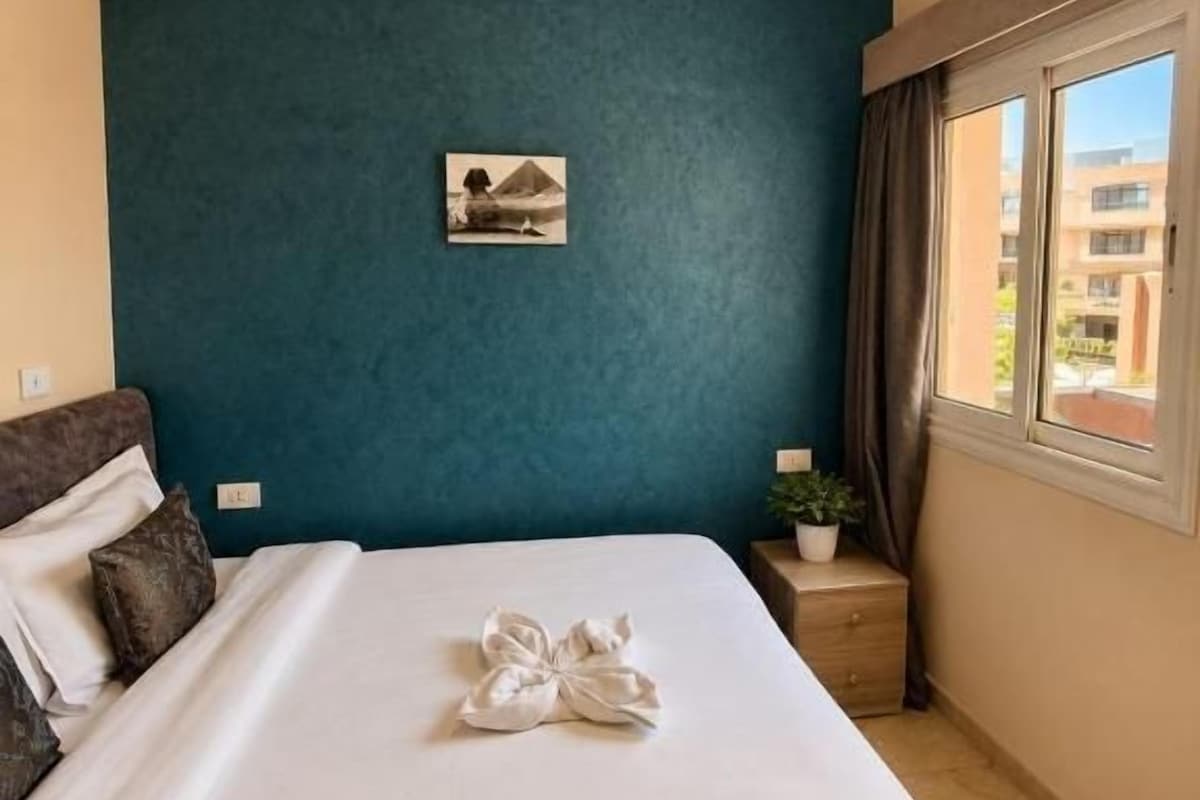 Ein Hotelzimmer mit einem Bett, einem Nachttisch, einem Fenster und einem an der Wand hängenden Bild.