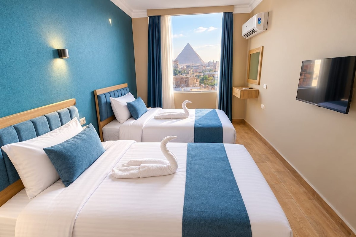 A hotel room with two beds, a TV, an air conditioning unit, and a view of a pyramid.