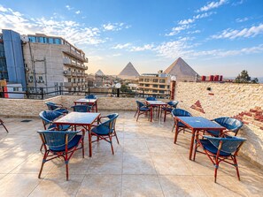 Una terraza al aire libre con mesas y sillas, un muro de piedra y vistas a pirámides al fondo.
