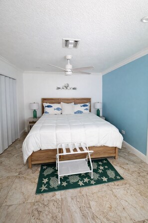 Une chambre à coucher comprenant un lit, un ventilateur de plafond, une table de chevet et un tapis vert orné de motifs représentant des tortues de mer et des étoiles.