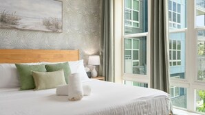 Hotellihuone, jossa on suuri sänky, sängynpääty, taulu seinällä ja näkymä rakennuksiin ikkunasta.