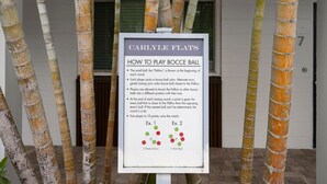 Skilt med instruktioner til petanque, placeret foran en bygning med bambuspiller.