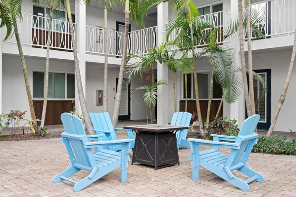 A courtyard with a table and four blue chairs, surrounded by palm trees and a building with balconies.