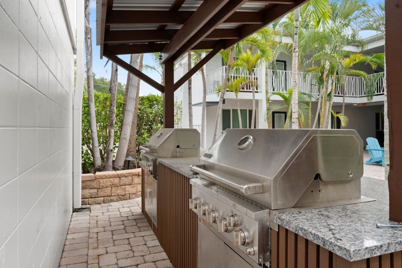 An outdoor barbecue area with two grills, a covered seating area, and a view of a building with balconies and palm trees.