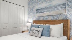 En pænt redt seng med en pude, der står 'velkommen HJEM', i et hotelværelse med tapet med mønster og et indrammet billede af en strand.