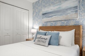 A neatly made bed with a pillow that reads 'welcome HOME' in a hotel room with a patterned wallpaper and a framed picture of a beach scene.