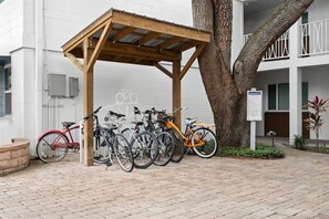 Pequeno estacionamento coberto para bicicletas com várias bicicletas estacionadas embaixo, uma árvore e um edifício com fachada branca.
