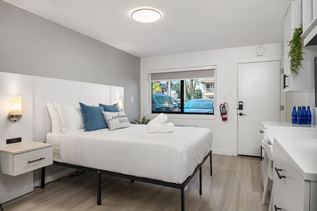 A modern hotel room with a bed, bedside table, and a window with a view of parked cars.