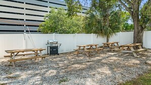 Un patio al aire libre con mesas de picnic y una parrilla, rodeado por una valla blanca y árboles.