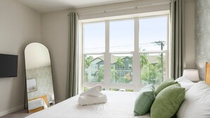 A hotel room with a large bed, a flat-screen TV mounted on the wall, a window with a view of buildings and trees, and a mirror with a leaf pattern. - Luxury Palm Retreat - HUGE 2BR 2BA near Downtown (Tampa)