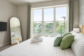 Ein Hotelzimmer mit einem großen Bett, einem am Wand montierten Flachbildfernseher, einem Fenster mit Blick auf Gebäude und Bäume sowie einem Spiegel mit Blattmuster.