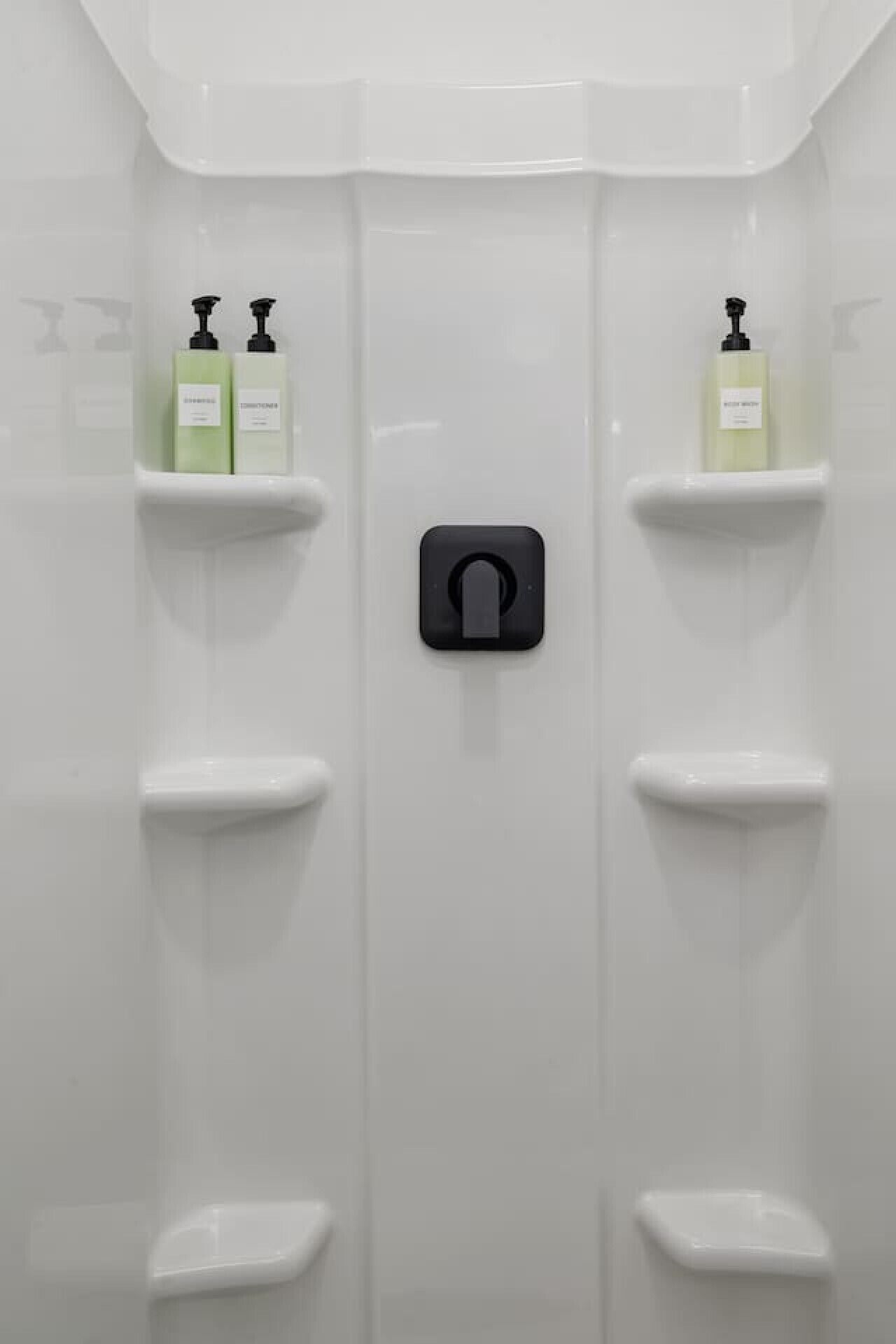 Un baño con pared de azulejos blancos, un dispensador de papel higiénico negro y dos estantes con botellas.