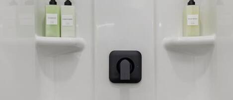 Un baño con pared de azulejos blancos, un dispensador de papel higiénico negro y dos estantes con botellas.
