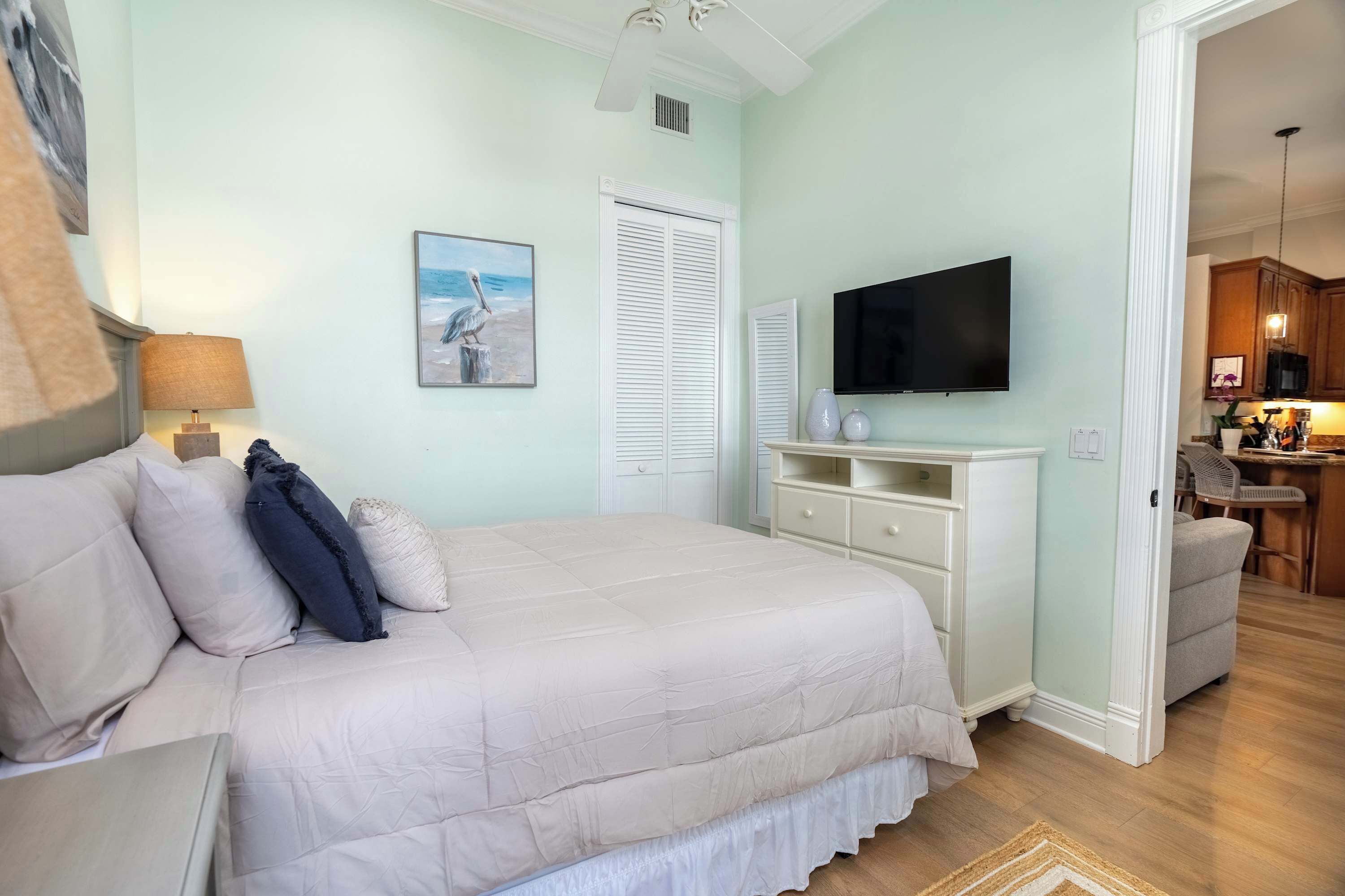 A bedroom with a large bed, a television, a painting, and a view into the kitchen.