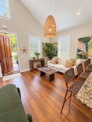 A spacious living room with a wooden floor, a large woven pendant light, a white sofa, a coffee table, and a dining area with wicker chairs and a granite table. - Tortugas Harbor Light Suite (Key West)