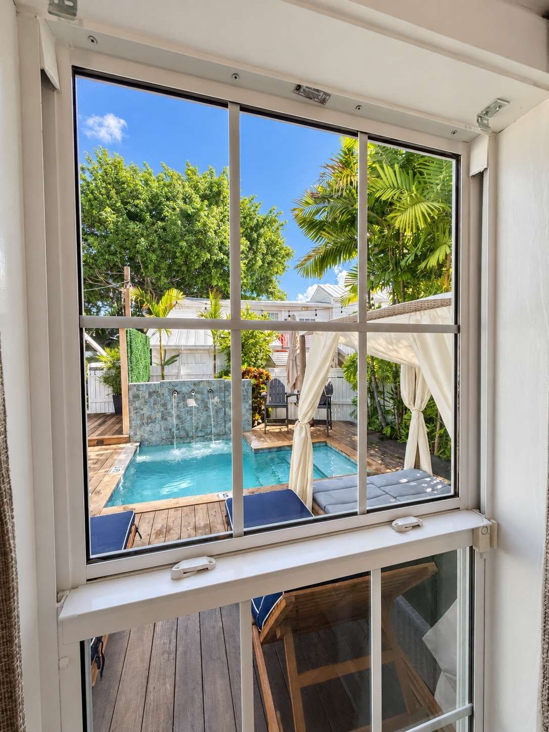 Blick durch ein Fenster in einen Garten mit Schwimmbad, Whirlpool und einer Holzterrasse.