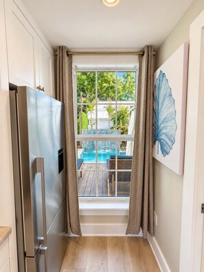 Une cuisine équipée d’un réfrigérateur en acier inoxydable, d’une fenêtre avec des rideaux et offrant une vue sur une piscine et du mobilier d’extérieur.