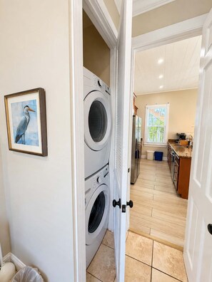 A laundry room with stacked washers and dryers, a framed picture on the wall, and a kitchen area in the background. - Alligator Reef Light Suite (Key West)