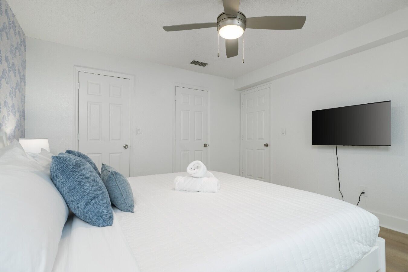 Una habitación de hotel con cama, ventilador de techo, televisor montado en la pared y una puerta.