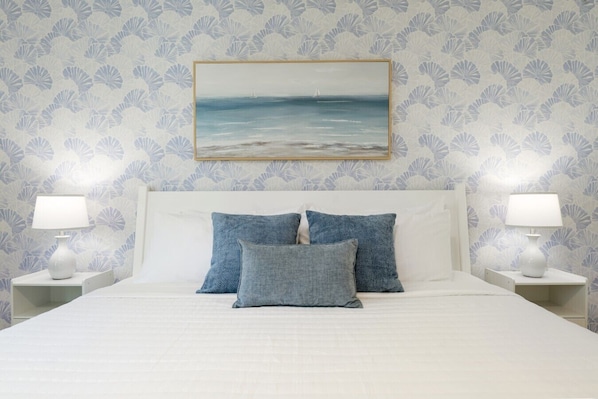 A bed with white bedding, two blue pillows, and a framed seascape painting above it.