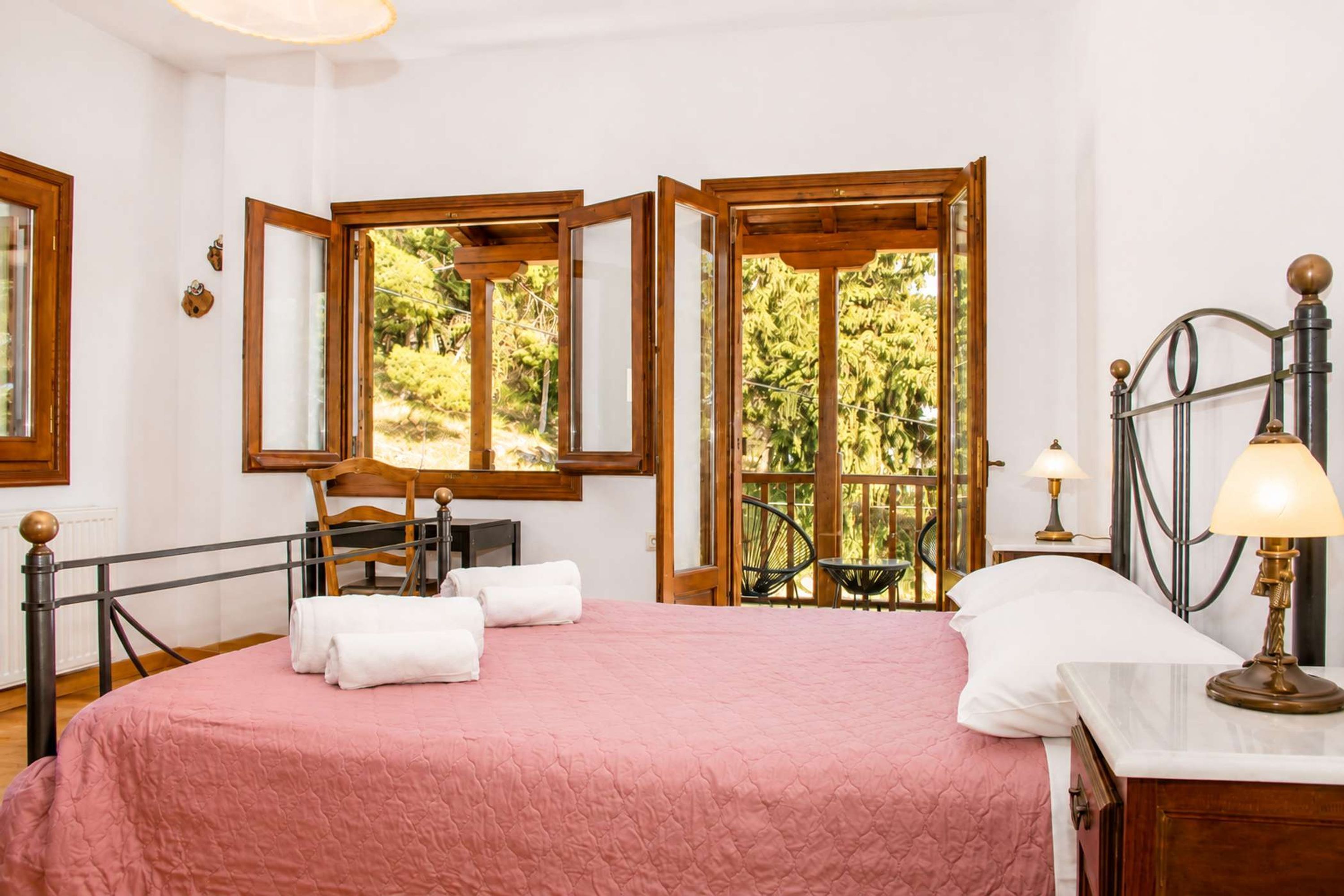 Une pièce comprenant un lit, un bureau et une chaise. Elle possède deux fenêtres à cadre en bois, et offre une vue sur la verdure à l’extérieur.