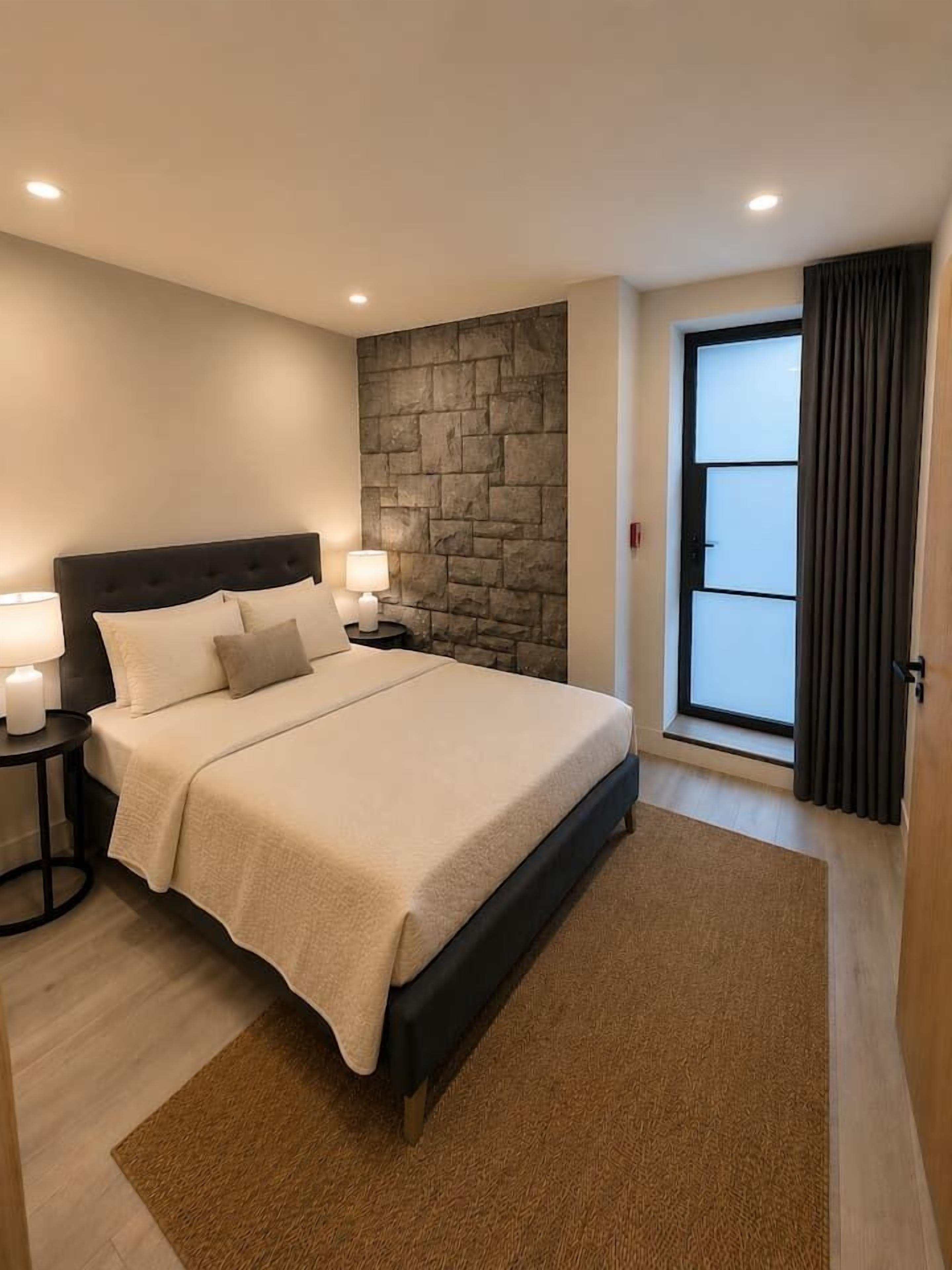 A bedroom with a bed, nightstands, a stone accent wall, a window with curtains, and a door.