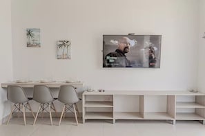 Ein minimalistisches Wohnzimmer mit einem an der Wand montierten Flachbildfernseher, einem Essbereich mit hellem Tisch und Stühlen sowie zwei gerahmten Kunstwerken an der Wand.