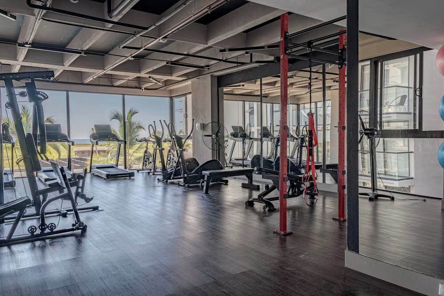 Uma academia com diversos equipamentos de exercícios, janelas amplas e vista para palmeiras.