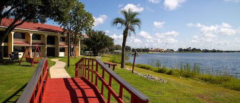 Un puente de madera que lleva a un edificio con techo rojo, una zona cubierta de césped y una masa de agua.