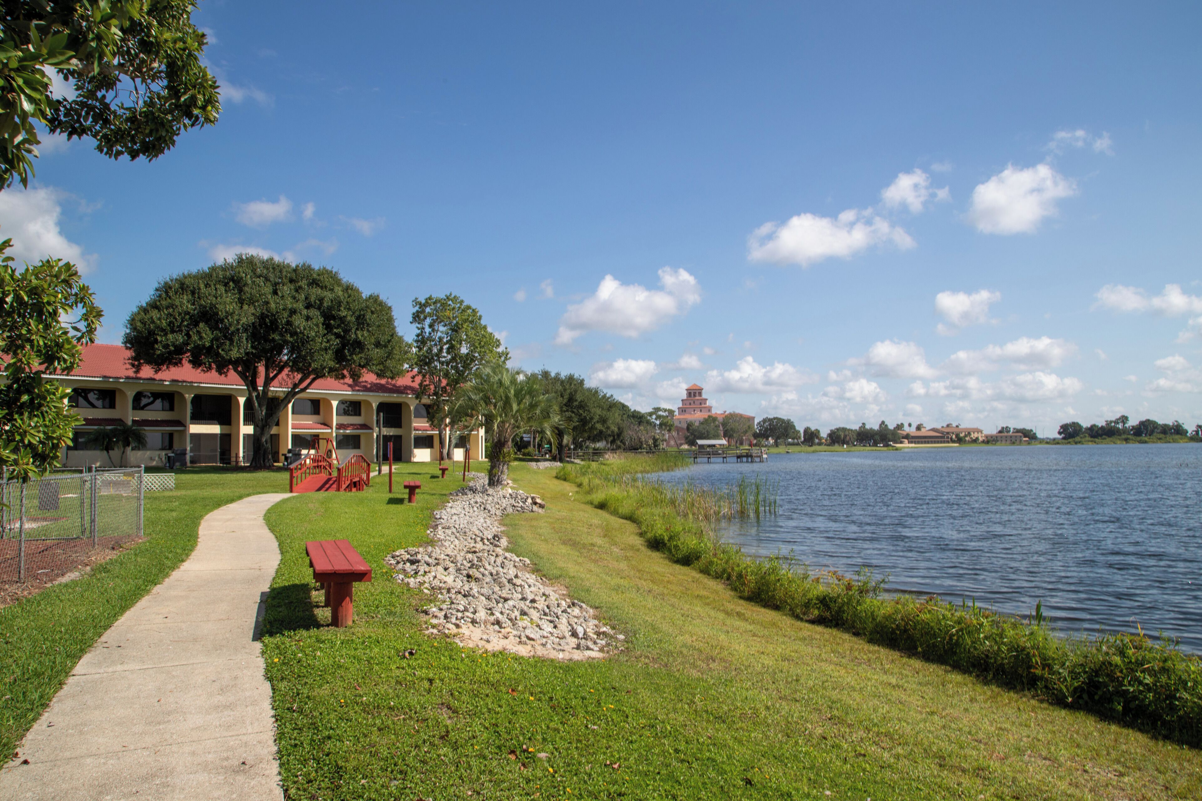 A lakeside walkway with a picnic table, a building with a red roof, and a tower structure across the water.