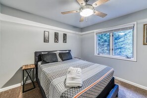 Quarto com cama, ventilador de teto, janela com vista para árvores e quadros nas paredes.