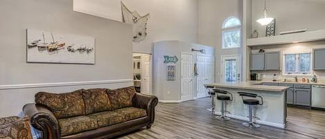A spacious living room with a large brown sofa, a kitchen area with a bar, and a dining area with a high ceiling and decorative wall art.