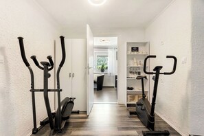 Ein Flur mit zwei Ergometer, einem Topf mit Pflanze und Blick in einen Raum mit Fenster und Regal mit Gegenständen.
