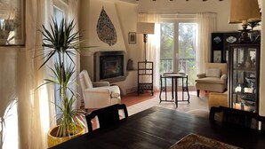 A cozy living room with a fireplace, a dining table, and a view of the outdoors through large windows.