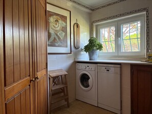 Eine kleine Küche mit Waschmaschine, einem Hocker, einer Topfpflanze und einem Fenster mit Blick auf Bäume.