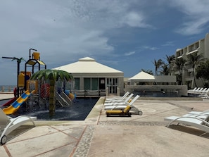 A pool area with a water play structure, lounge chairs, and a building with a white roof. - Three Bedroom Beachfront Villa - Private Heated Pool & Beach Access (Cancun)