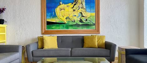 A living room with a sofa, a glass coffee table, and a framed painting on the wall.