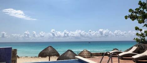 A pool with a view of the beach and ocean, lounge chairs, and thatched umbrellas.