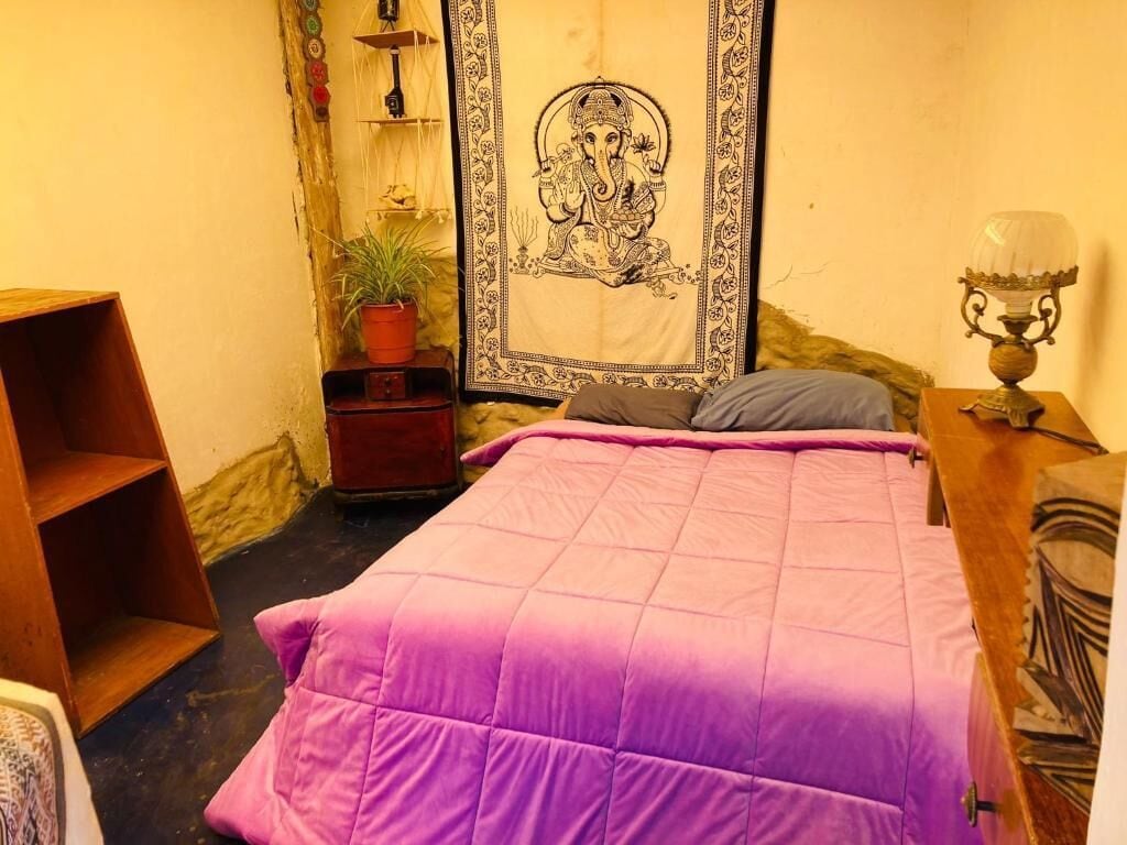 Un dormitorio con cama, mesita de noche, lámpara, estante con adornos y un tapiz.