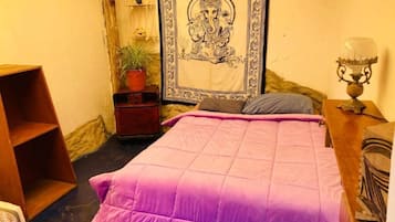 Un dormitorio con cama, mesita de noche, lámpara, estante con adornos y un tapiz.