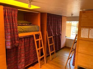 A dormitory room with bunk beds, a wooden ladder, a window with curtains, and a bulletin board. - Kamanti Hostel (Pisac)