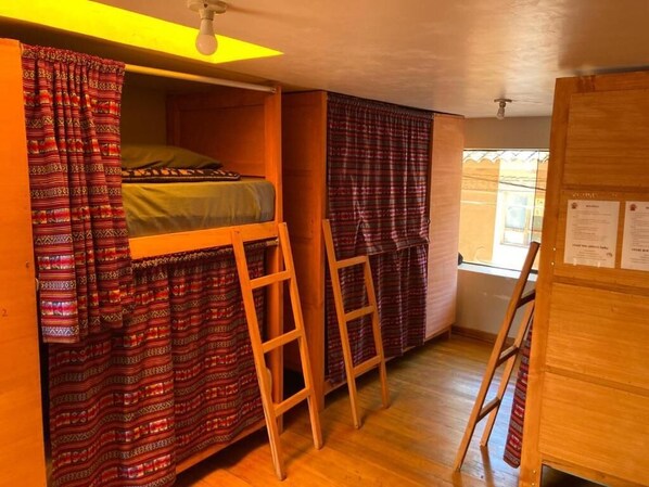A dormitory room with bunk beds, a wooden ladder, a window with curtains, and a bulletin board.