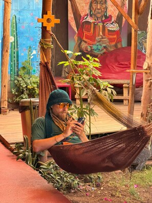 A person sitting in a hammock outdoors with a mobile phone, surrounded by plants and a mural in the background. - Kamanti Hostel (Pisac)