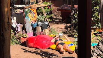 Una persona acostada sobre una esterilla al aire libre, rodeada de plantas y un edificio con un letrero.