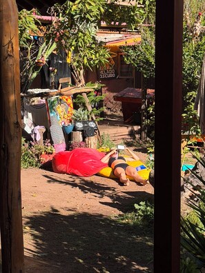 A person lying on a mat outdoors, surrounded by plants and a building with a sign. - Kamanti Hostel (Pisac)
