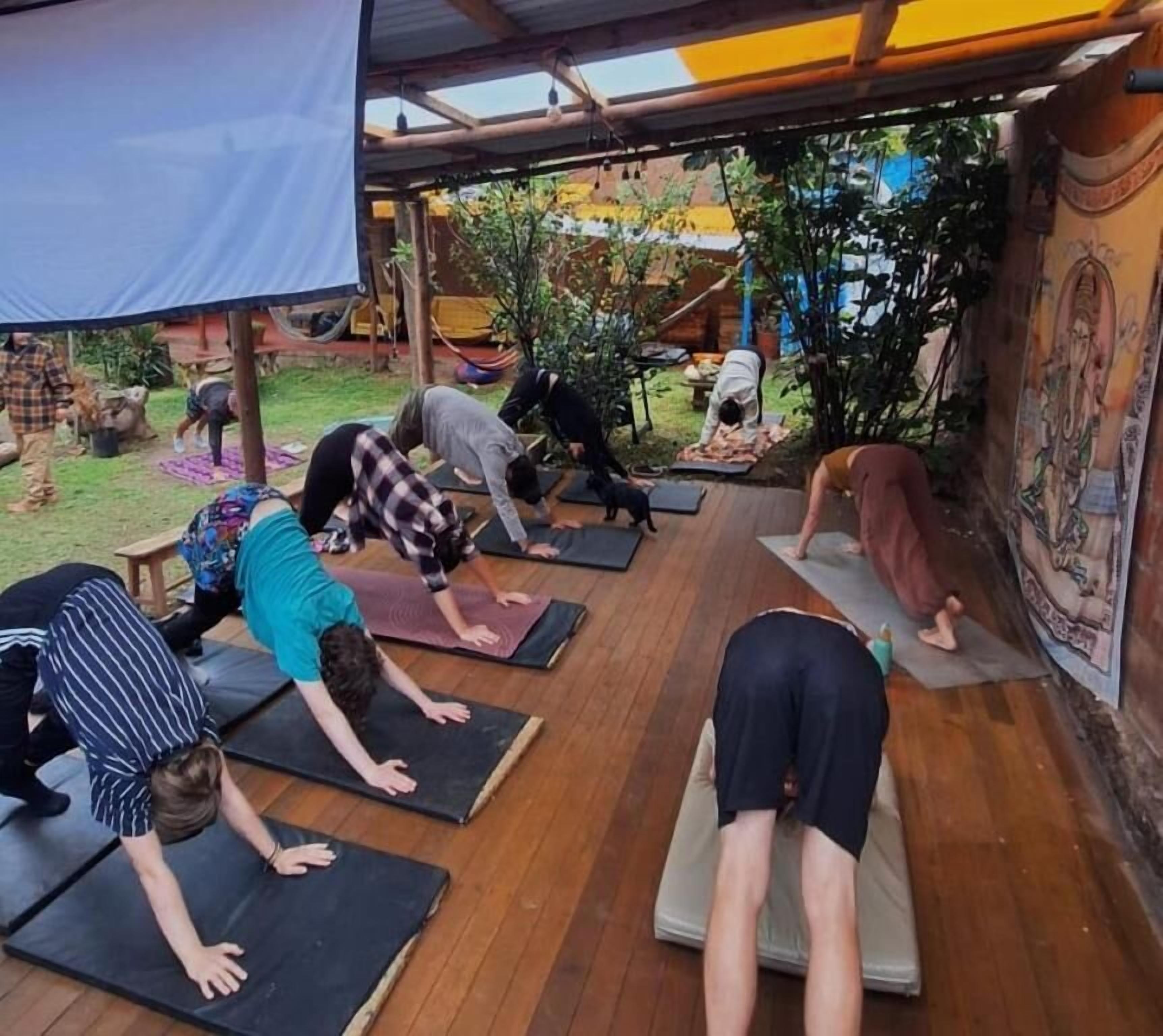 Un grupo de personas practicando yoga sobre colchonetas en un espacio al aire libre con suelo de madera y una zona cubierta.