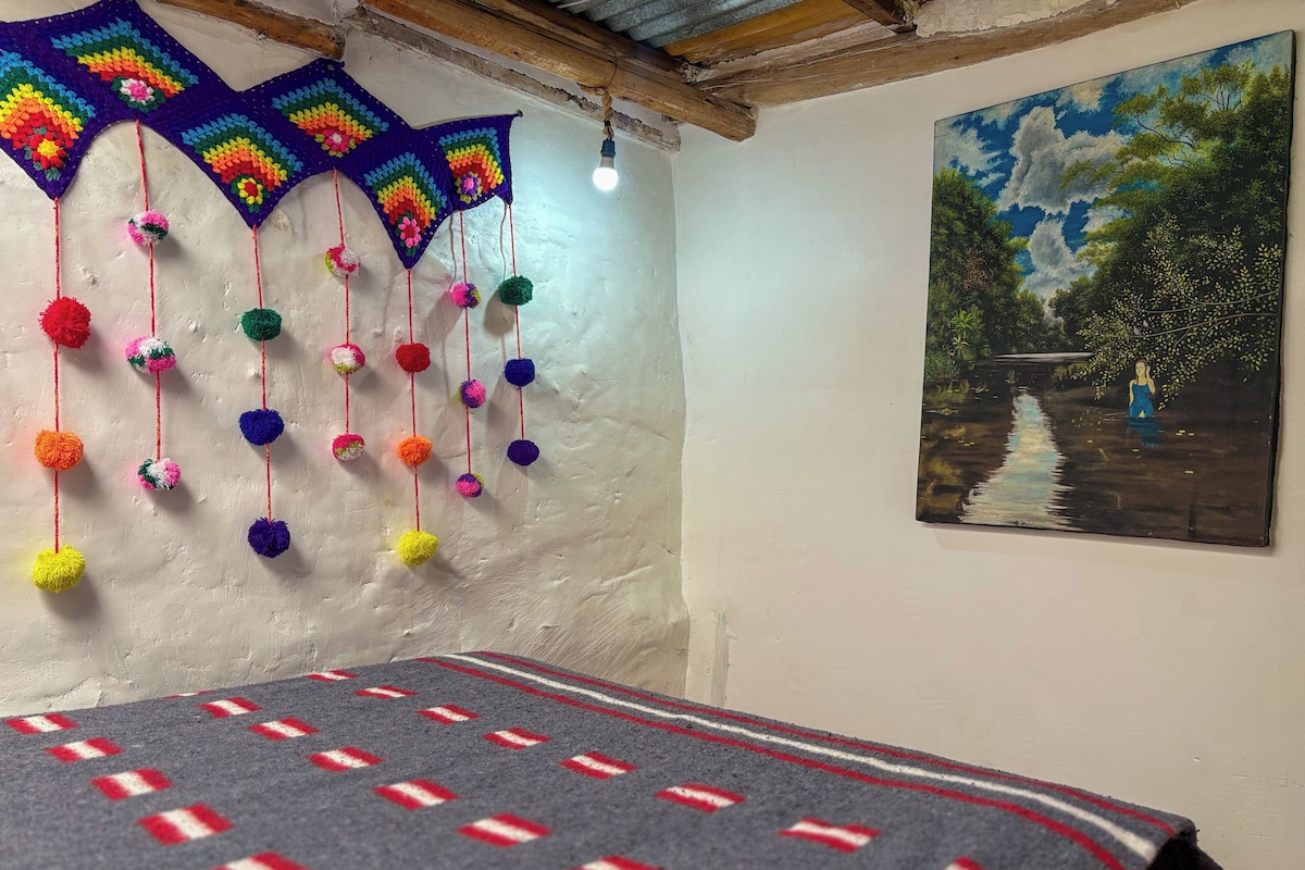 A room with a bed, a colorful crocheted wall hanging, and a painting.